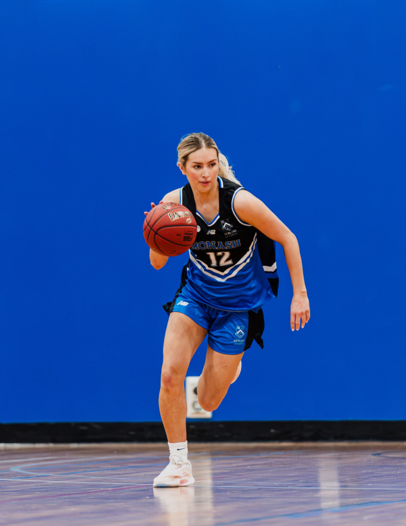 Female basketball player in a blue jersey (#12) dribbling toward the court against a solid blue background.