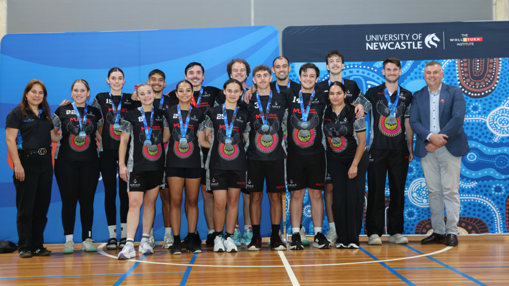 Group of athletes in matching black uniforms with medals around their necks, posing for a team photo in a gym with a University of Newcastle backdrop.