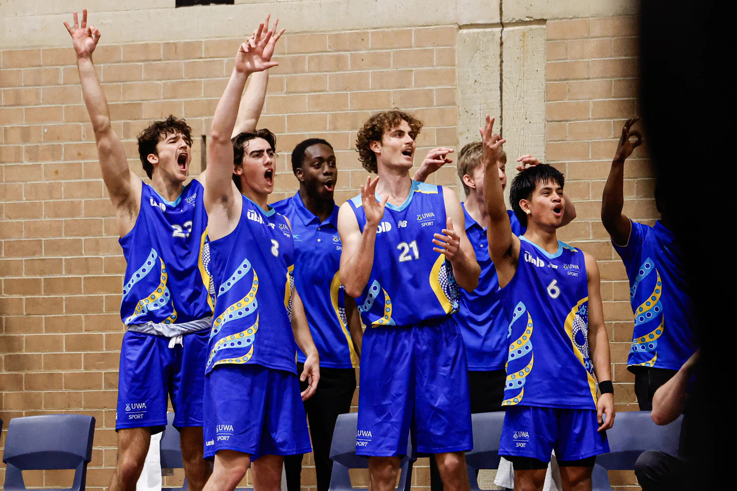 Group of young men in blue uniforms cheering with raised arms in a gym against a brick wall onlookers seated nearby.