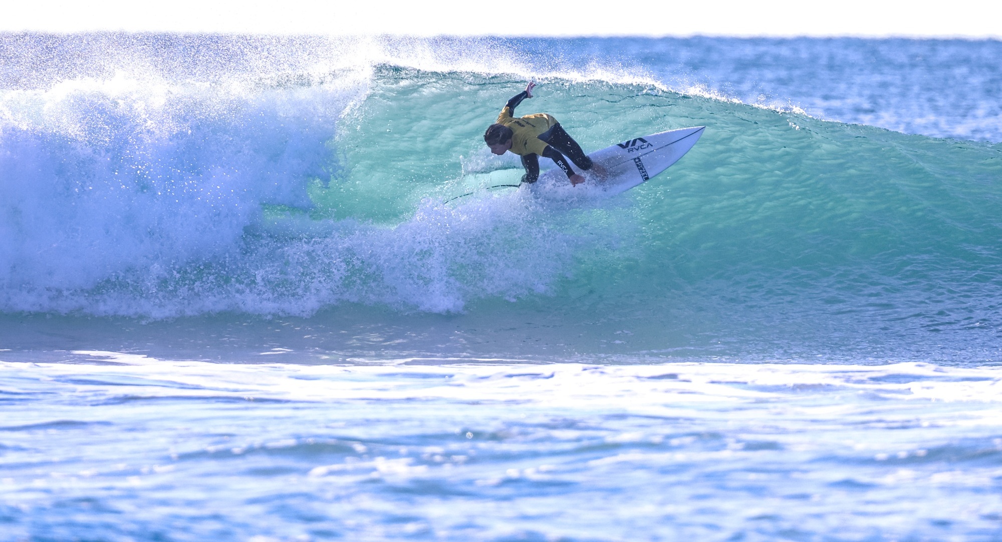 Surfer in a yellow shirt riding a green-blue wave on a white surfboard near the curl.