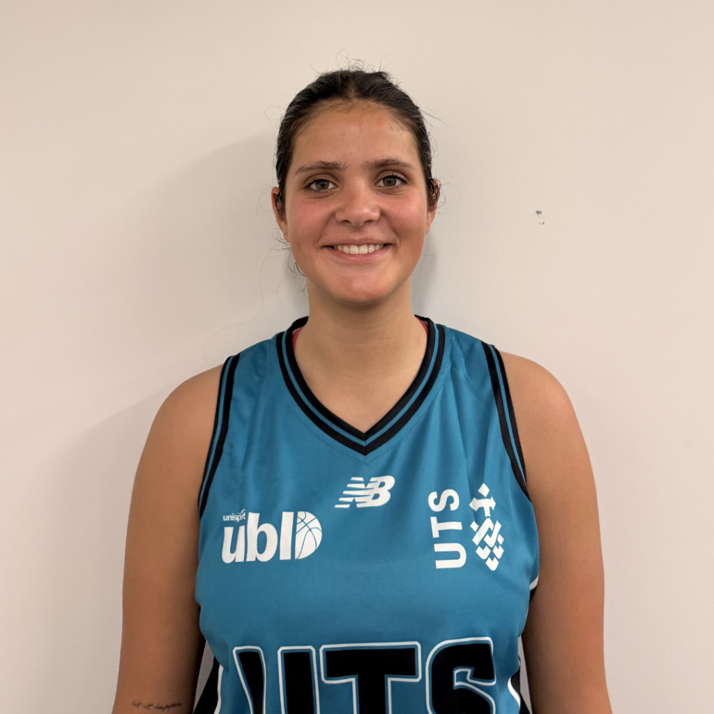 Portrait of a young woman in a teal basketball jersey, smiling, standing against a plain light wall.