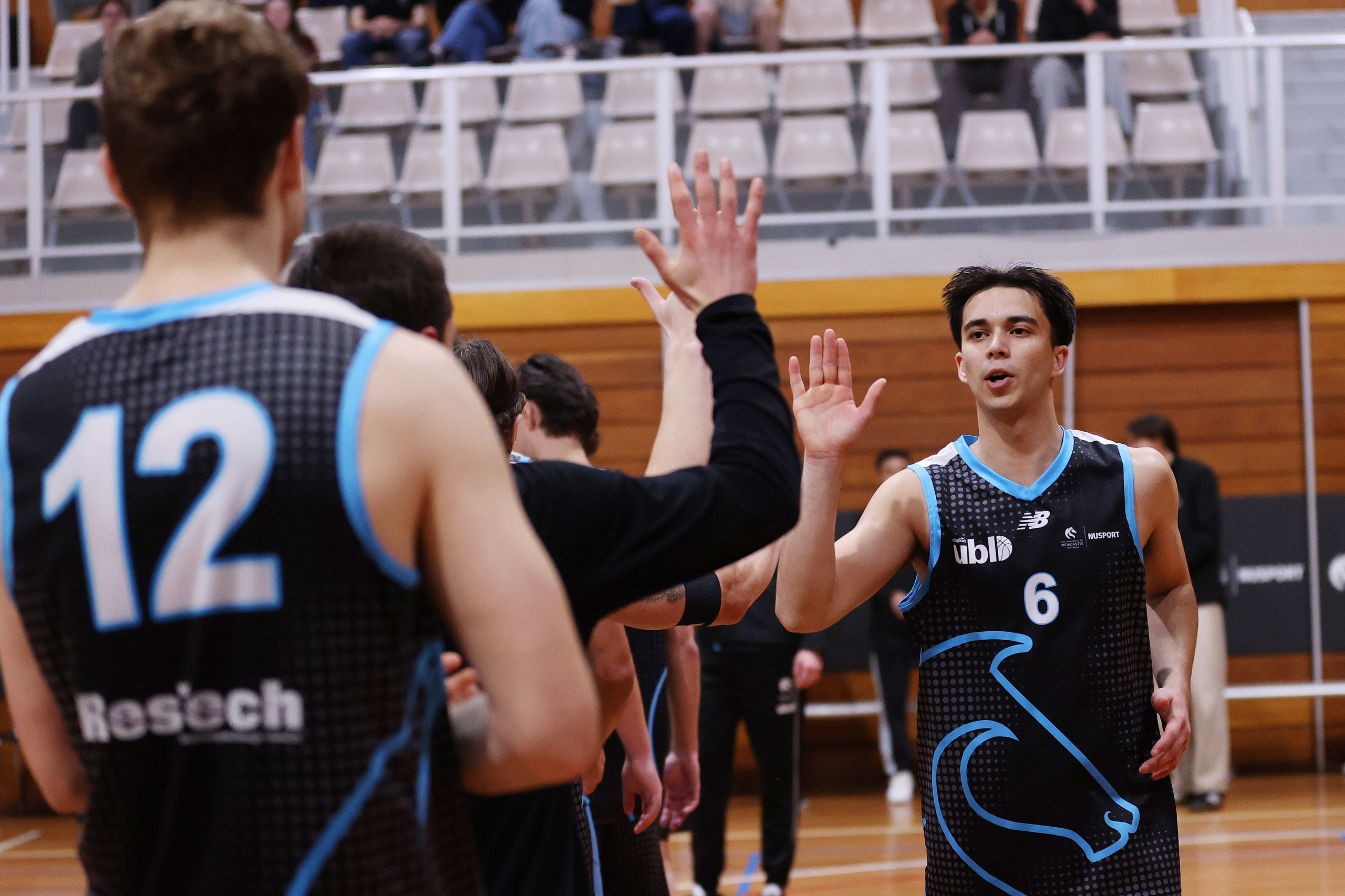 Basketball teammates in black and blue uniforms high-fiving in a gym with wooden bleachers in the background, number 6 visible on one player.