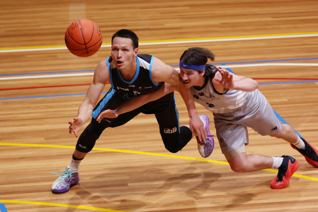 Two male basketball players sprint for a loose ball on a wooden indoor court as the ball flies between them.