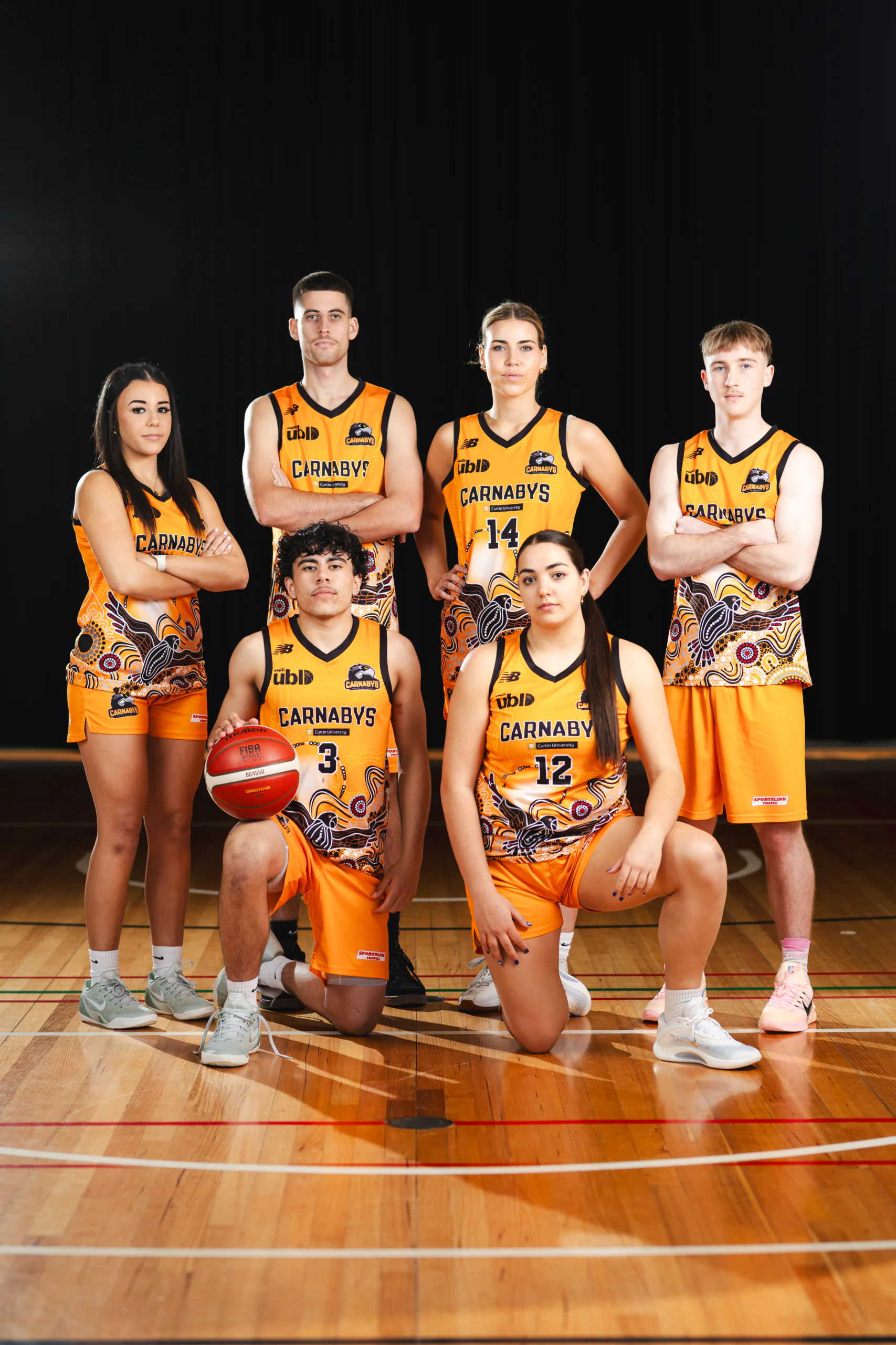 Six basketball players in orange Carnabys uniforms pose on a gym floor, two kneeling in front and four standing behind them.