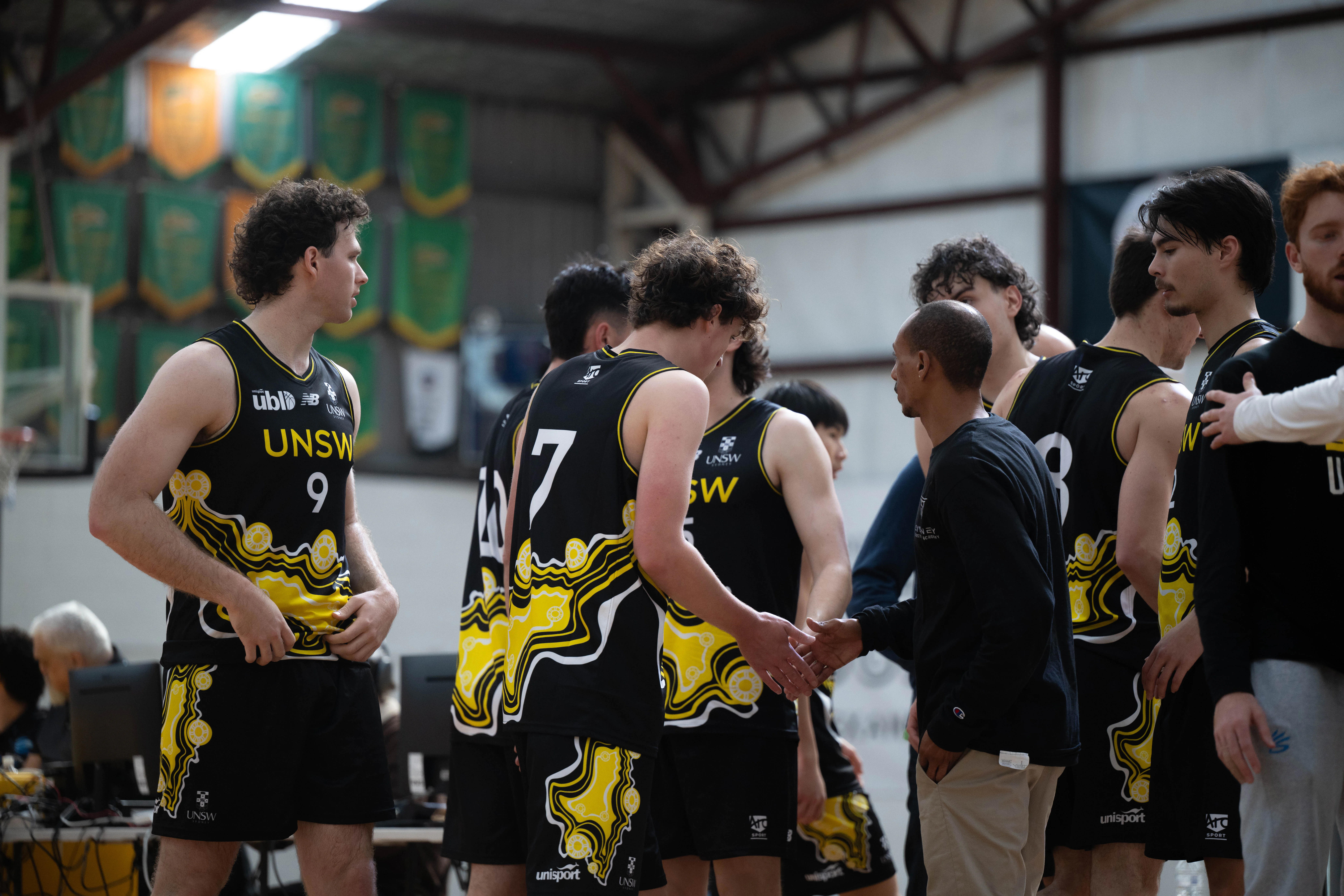 Basketball players in black and yellow uniforms shake hands after a game in a gymnasium.