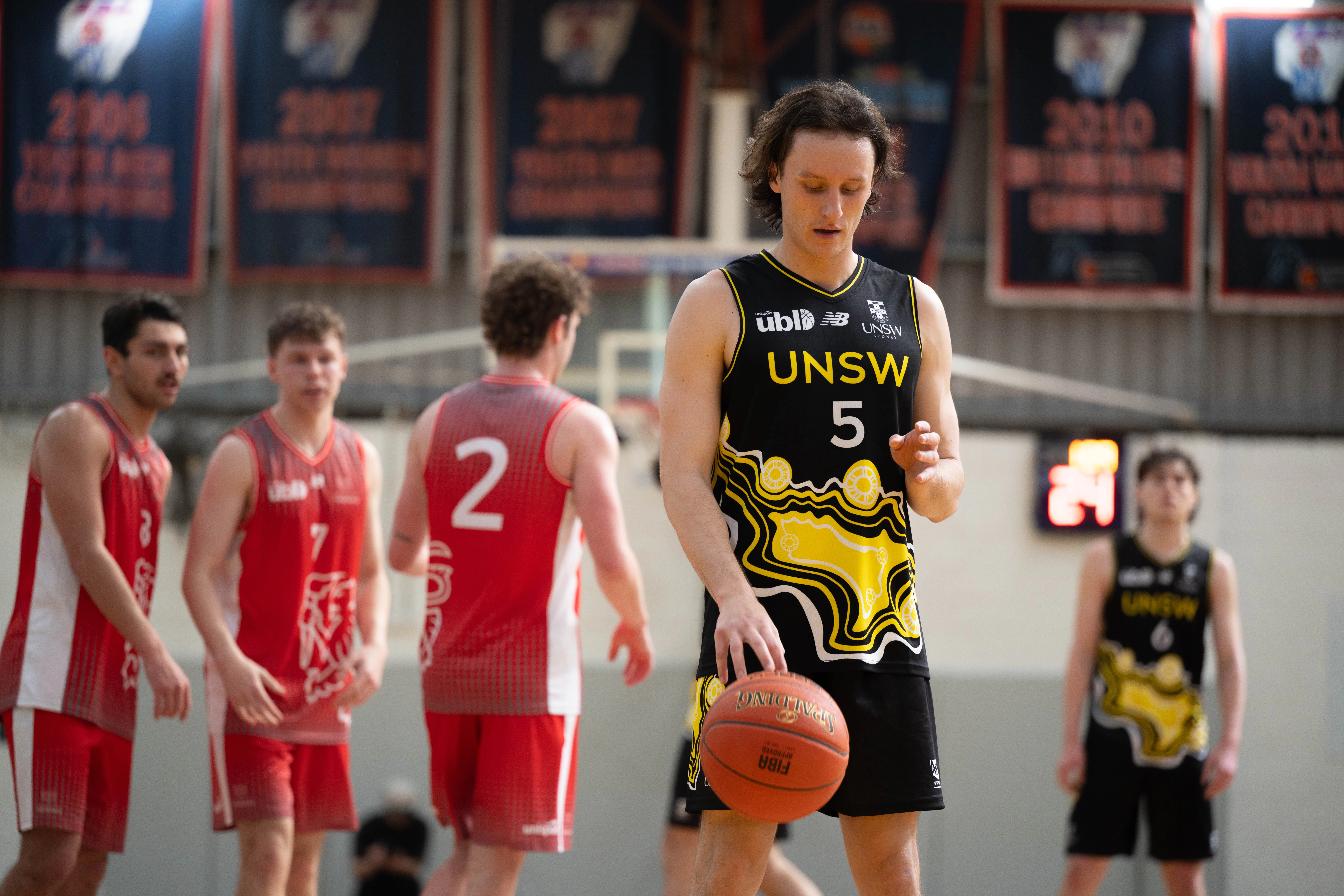 Basketball player in a black UNSW jersey with number 5 dribbles the ball on an indoor court, with red-uniform teammates in the background.