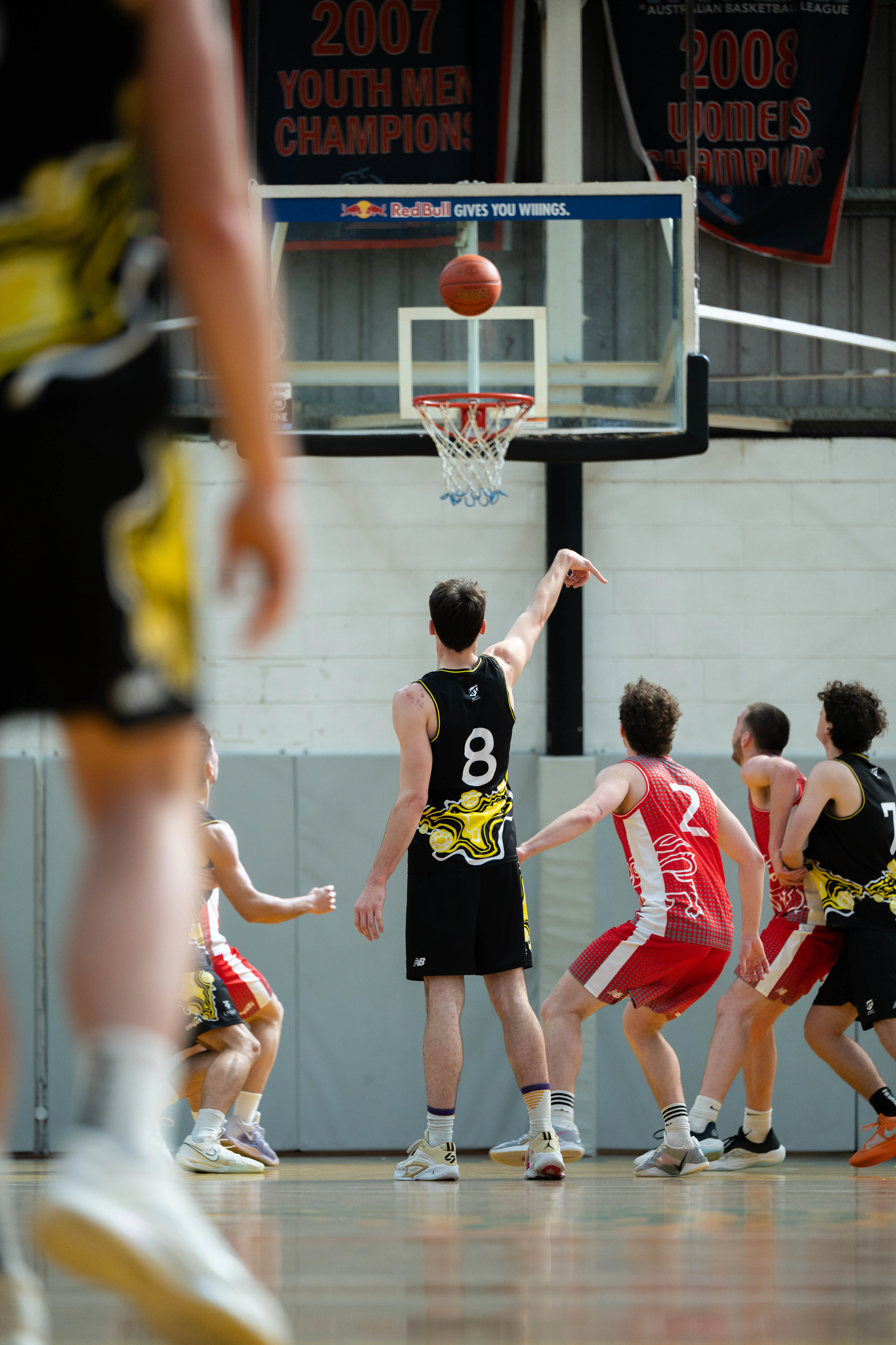 Basketball game in a gym; a player in a black jersey (#8) points toward the ball near the hoop while others defend.