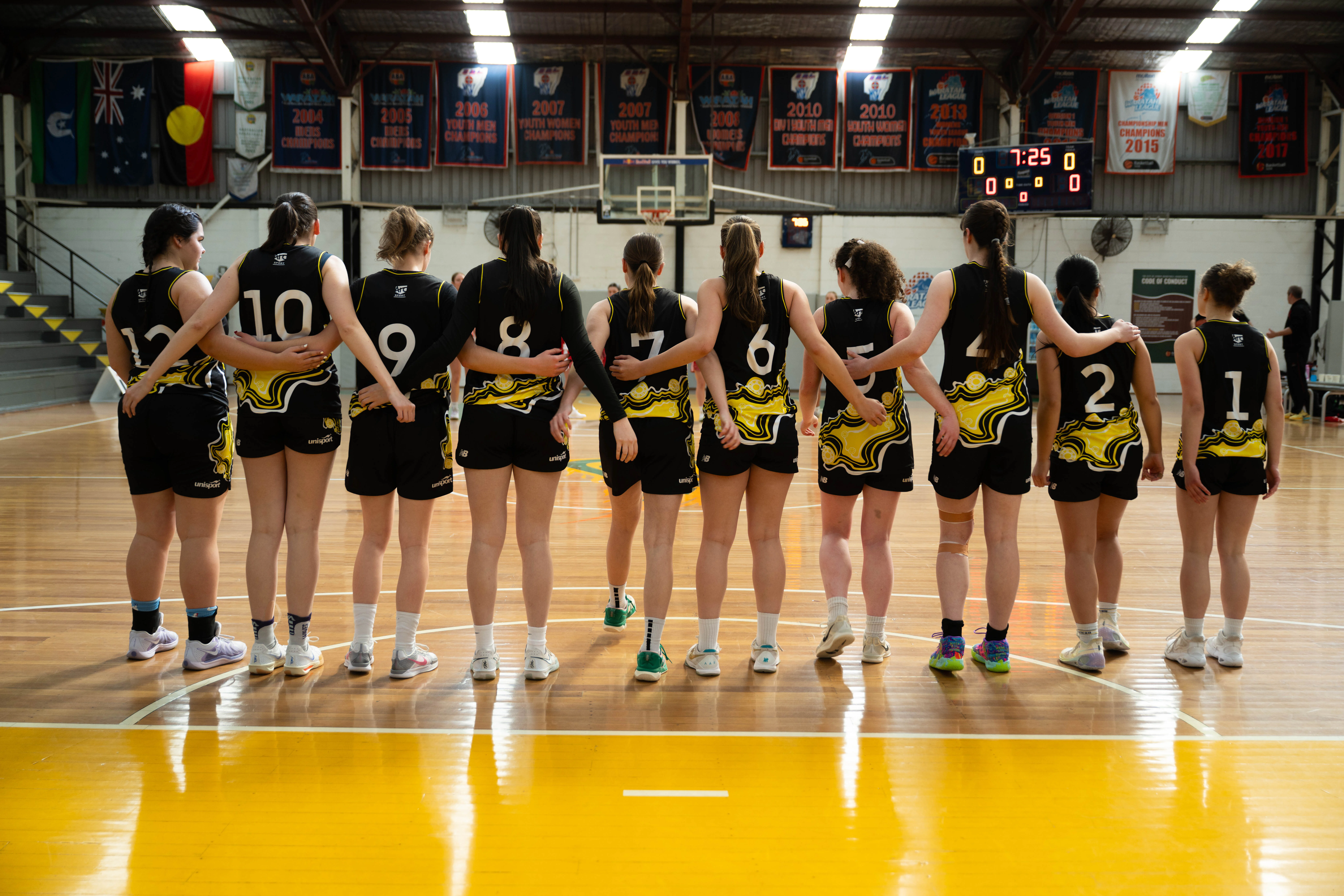 Girls basketball team huddling on a polished gym floor, wearing black and yellow uniforms with numbers on their backs.