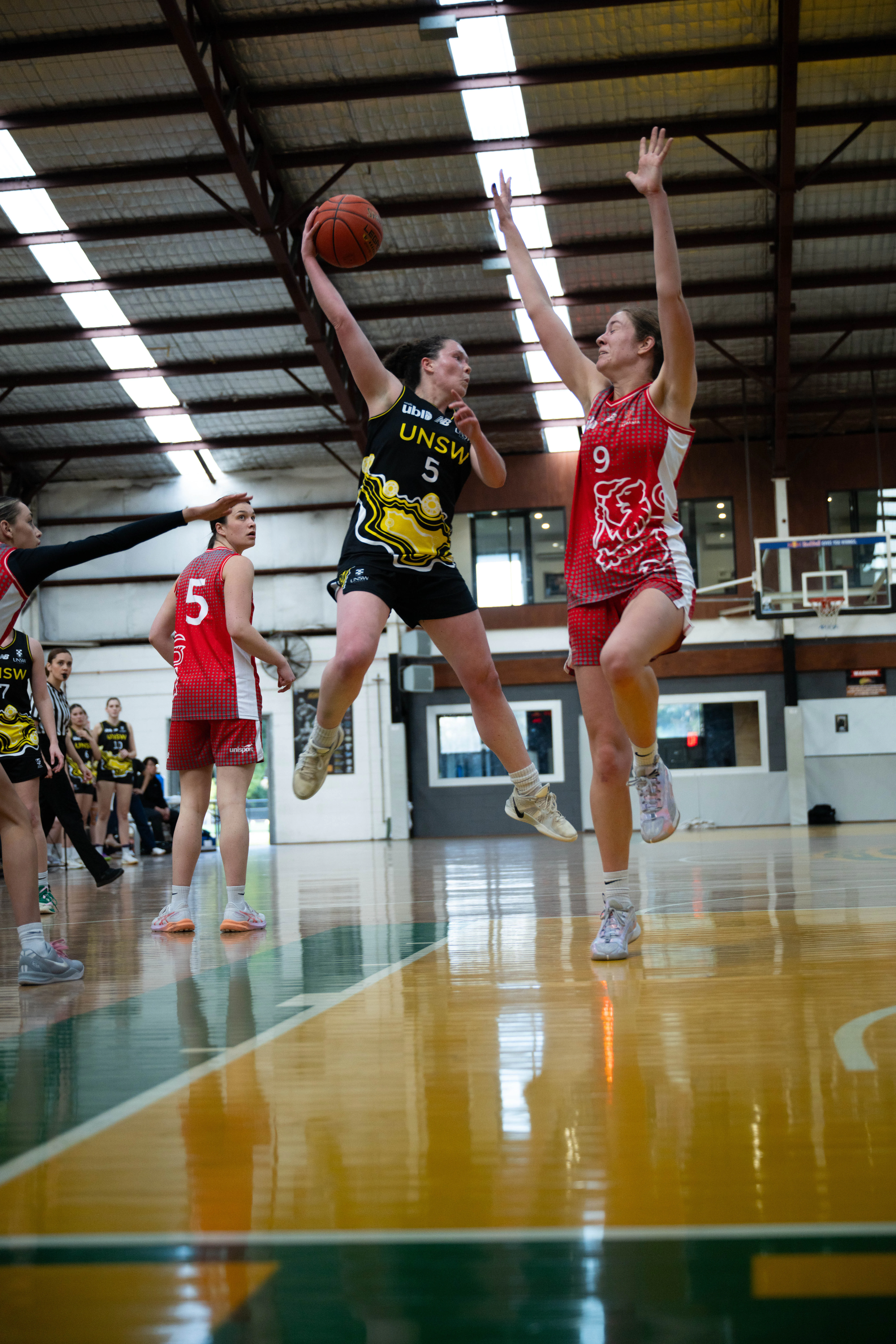 Two female basketball players leap for the ball in an indoor gym, one in a black UNSW uniform and the other in red, with teammates and a basketball hoop in the background.