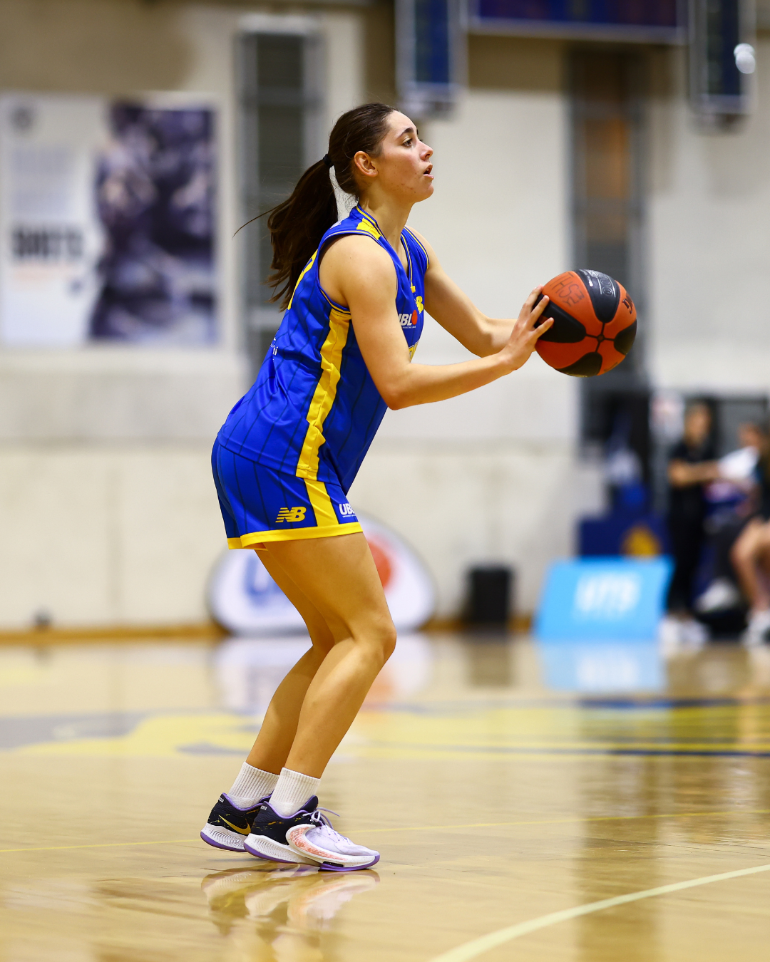 Female basketball player in a blue and yellow uniform holding a basketball, ready to pass or shoot on an indoor court.