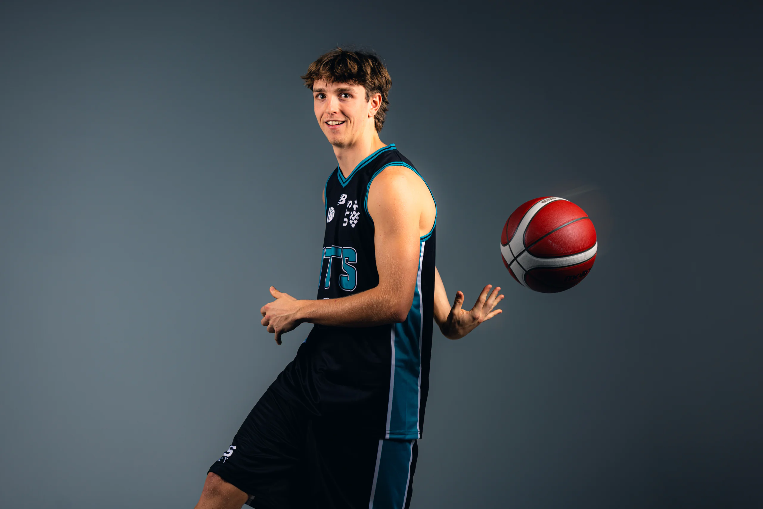 Male basketball player in teal and black uniform spins a red basketball against a gray studio backdrop, smiling at the camera.