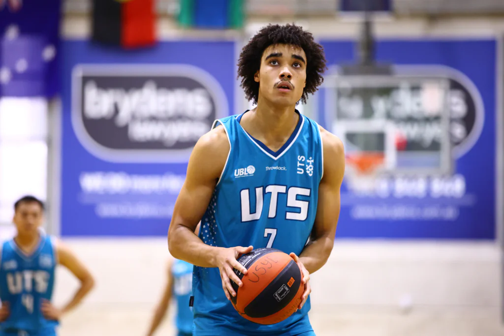 Young male basketball player in a blue UTES jersey holding a basketball on an indoor court, looking upward as if preparing to shoot or pass.