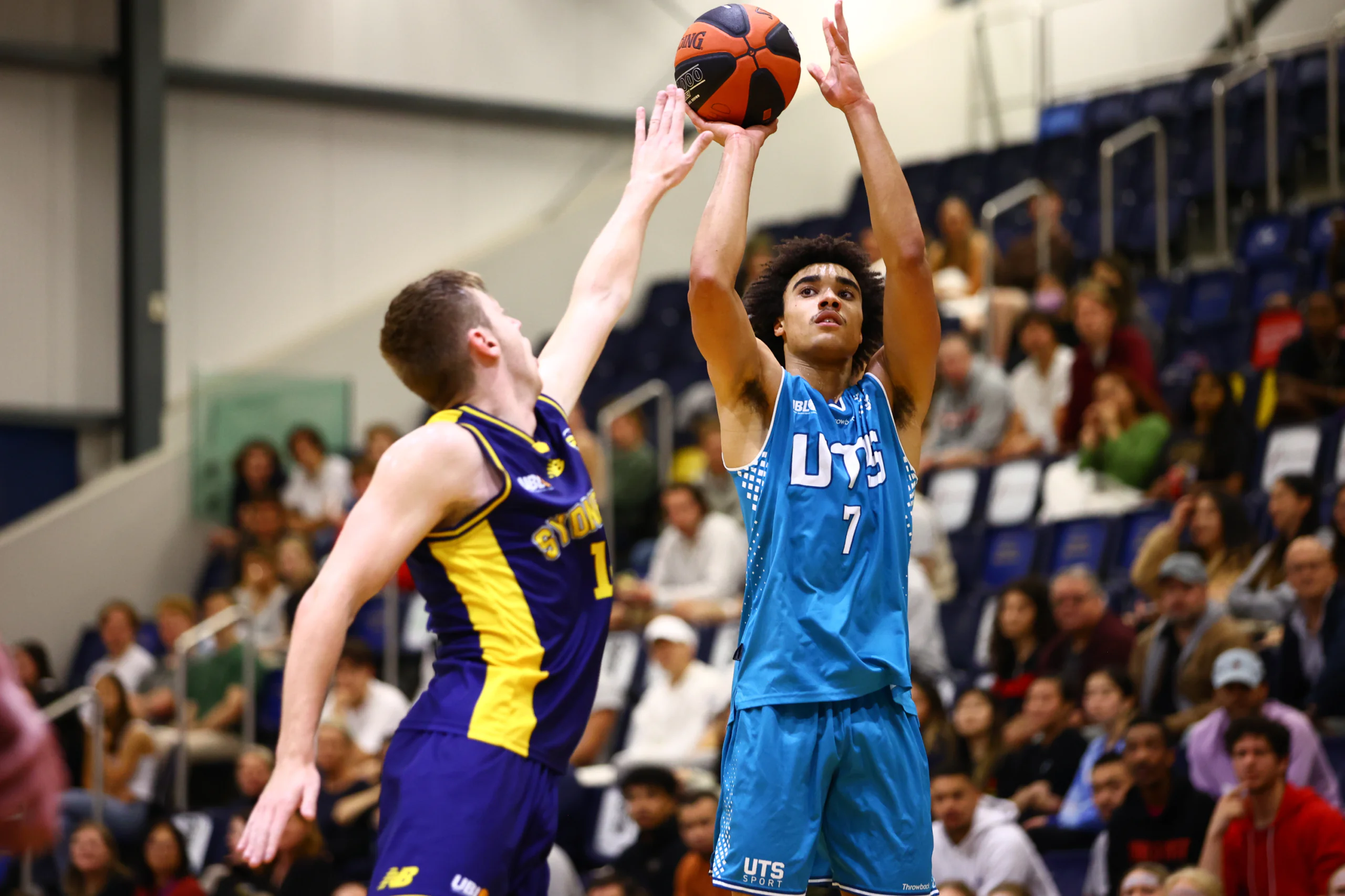 Two basketball players jump for the ball during a game; one in blue jersey reaching up against a defender in purple-yellow. Crowd in stands blurred in background.