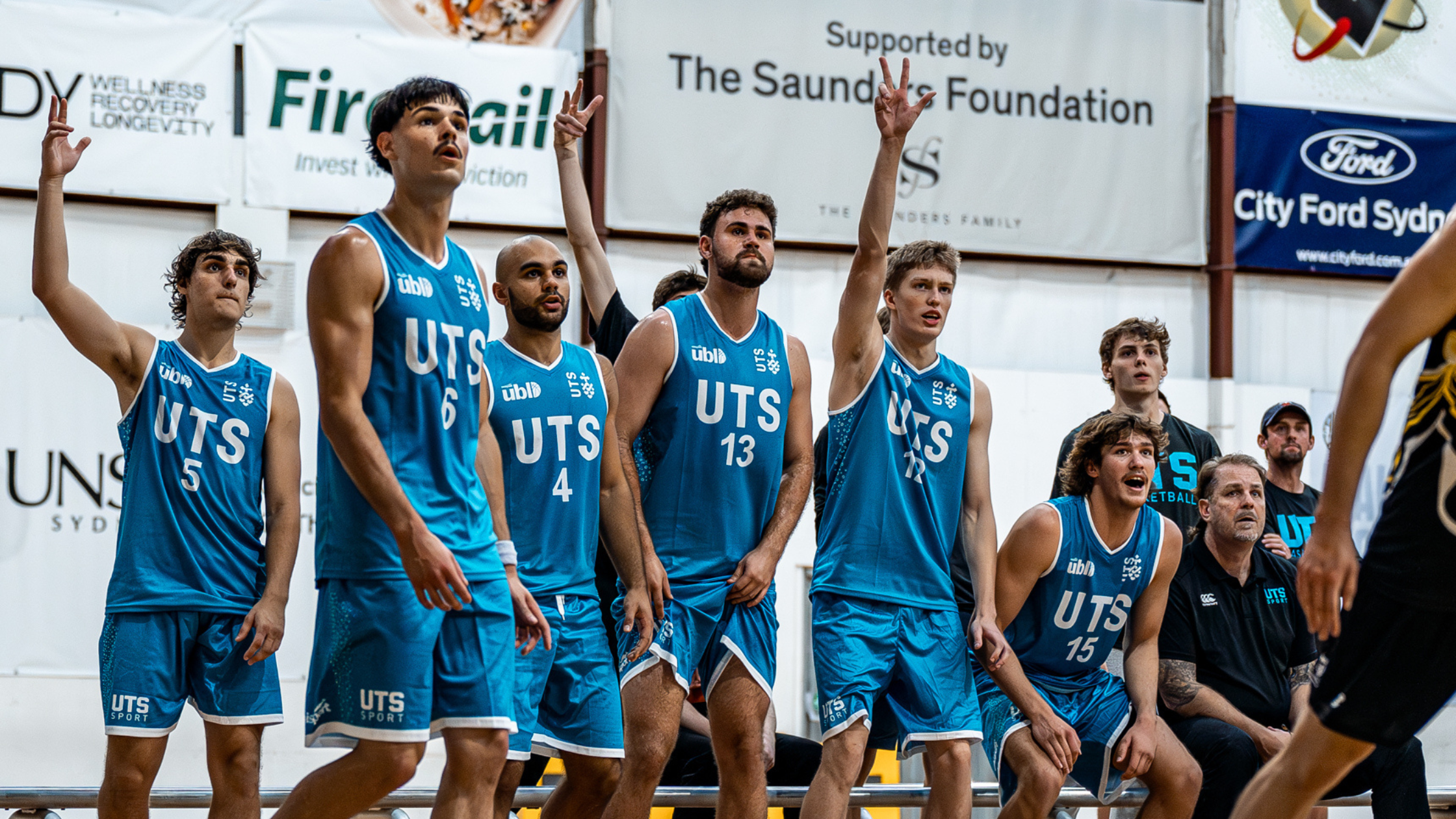 Group of young basketball players in blue UTS uniforms standing on an indoor court, some raising hands in a moment of team signaling.