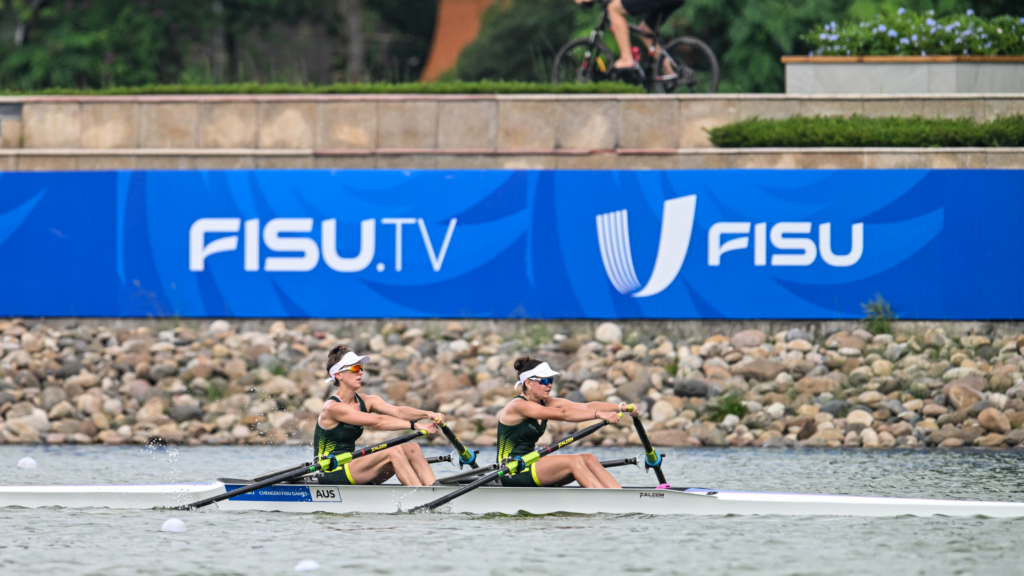Two female rowers race a double scull on a river, with a large blue FISU banner in the background.