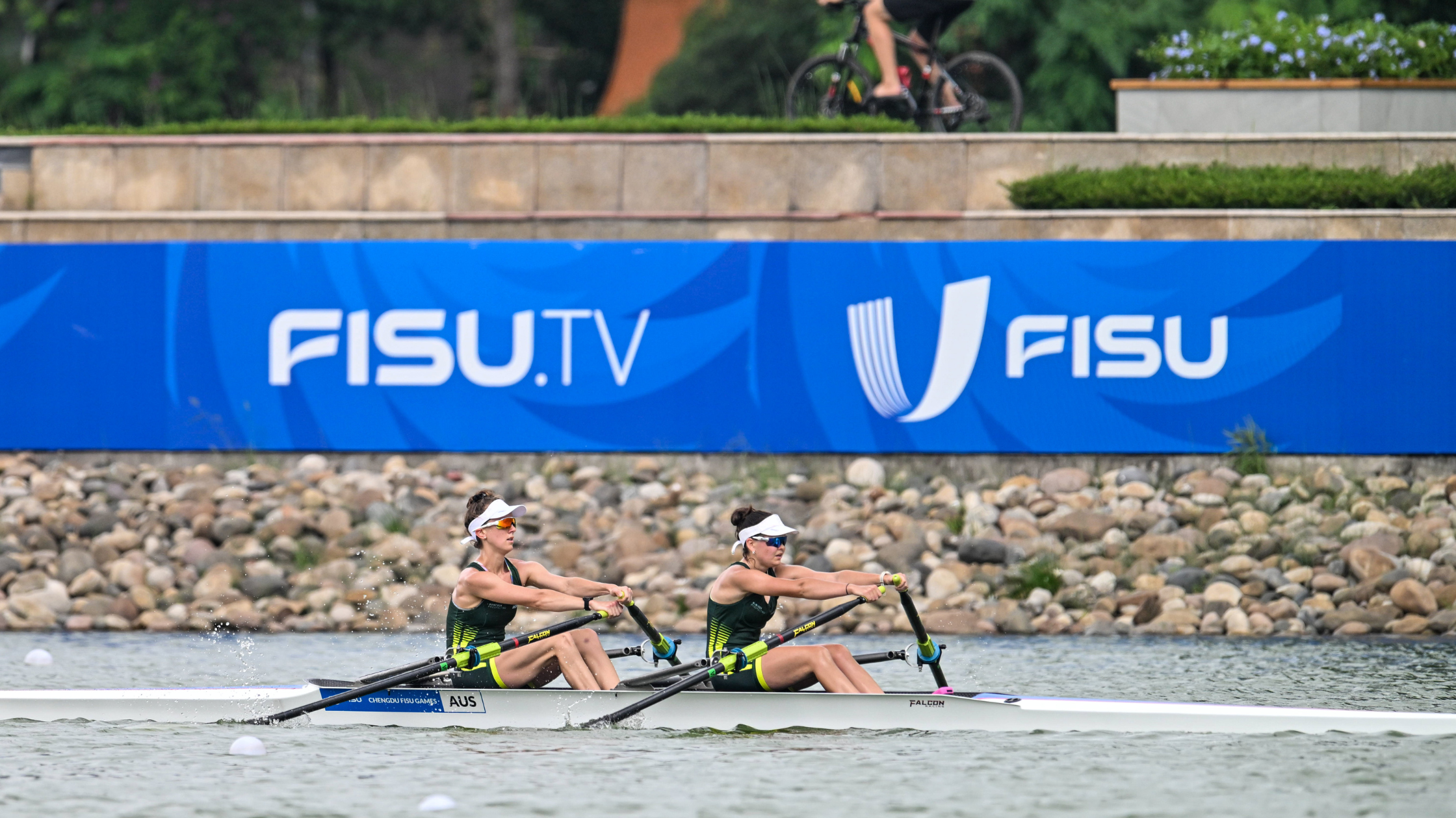 Two female rowers race a double scull on a river, with a large blue FISU banner in the background.