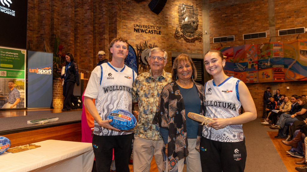 Group of two athletes in matching uniforms posing with an older couple in a university event hall, brick walls and colorful mural in the background.