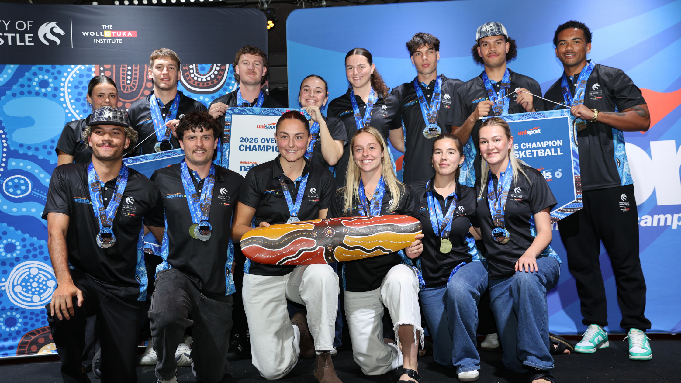 Group of athletes in black uniforms with medals posing on stage after winning a championship.