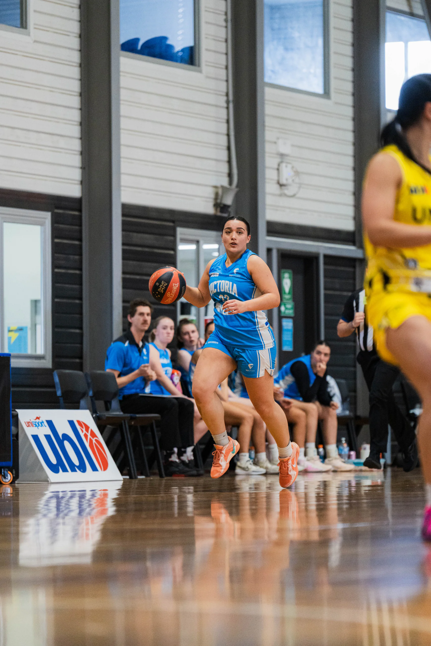 Female basketball player in blue uniform dribbles the ball on an indoor court, with teammates and spectators in the background.
