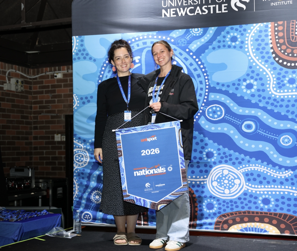 Two women stand on a stage posing for a photo, wearing lanyards; one holds a blue Indigenous Nationals bag in front of a blue patterned backdrop.