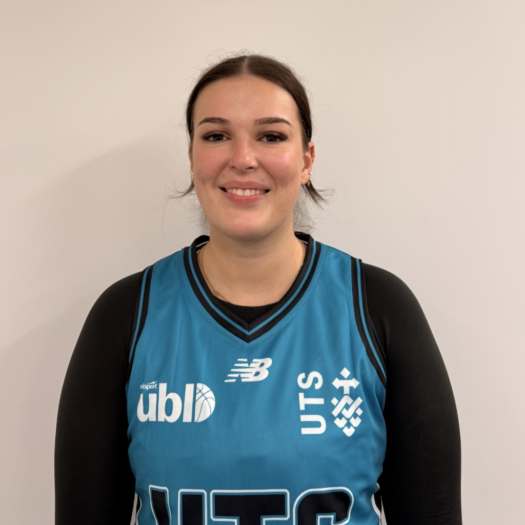 Smiling woman in a teal basketball jersey posing for a portrait against a light wall, facing the camera.