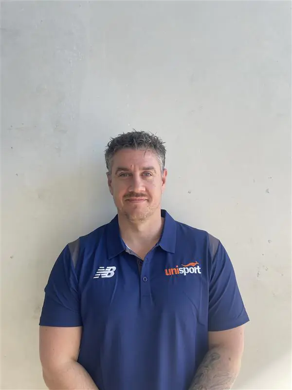 Portrait of a man in a navy polo with NB and UniSport logos, standing against a pale wall.