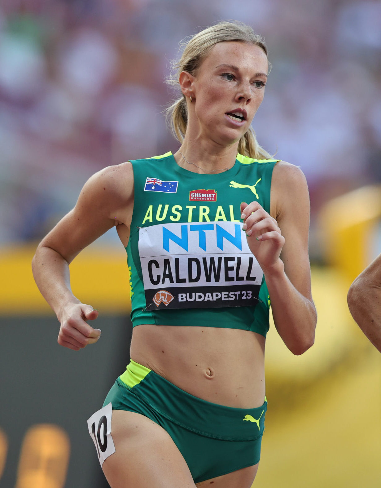 Australian female sprinter in green kit running, bib says CALDWELL at Budapest 23, focused expression
