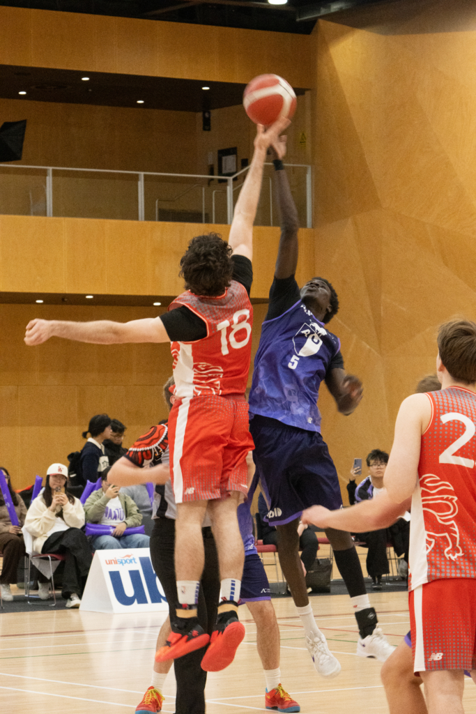 Two basketball players leap for the ball in mid-air during a game, one in red uniform and the other in blue, with spectators watching from the bleachers in a gymnasium.