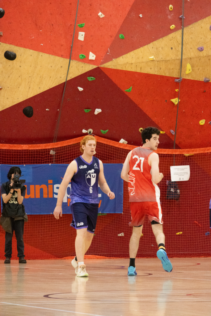 Two basketball players run on an indoor court; one in a blue uniform (number 6) and one in a red uniform (number 21), with a photographer on the left and a red safety net behind them.