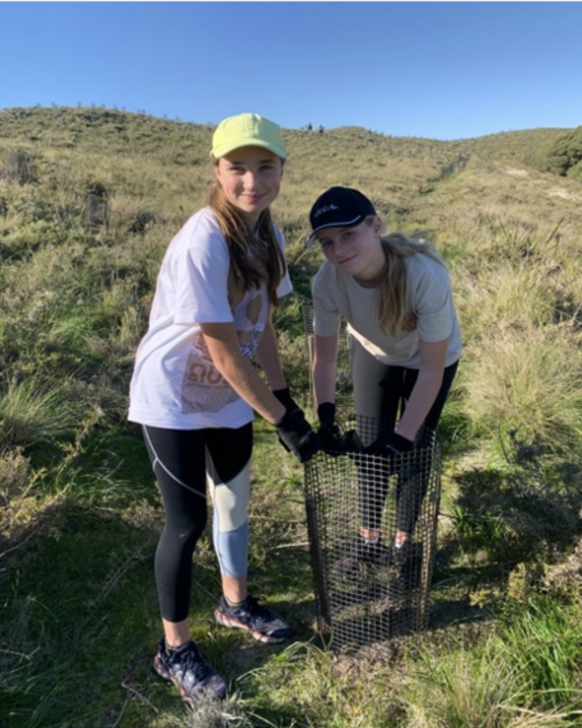 Two young women wearing gloves and caps stand on a sunny grassy hill, bending over a metal wire-mesh basket.