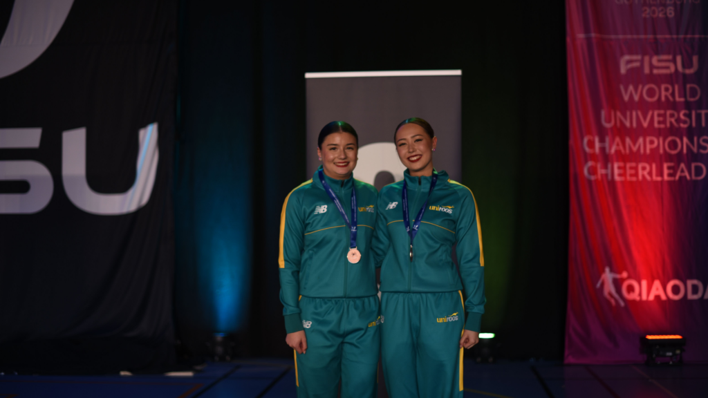 Two athletes in teal tracksuits with medals around their necks smile on stage after an awards ceremony.