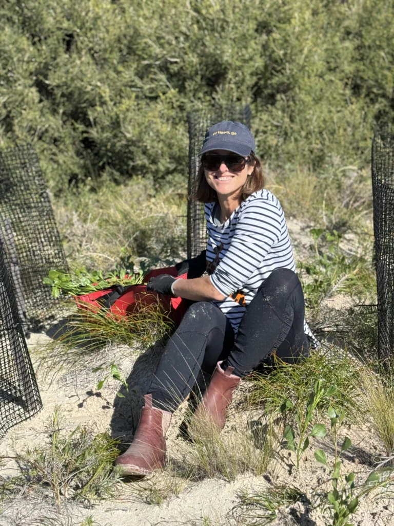 Smiling woman in a striped shirt and cap sits in a sandy planting bed with a red tote, gloves, and protective netting nearby.