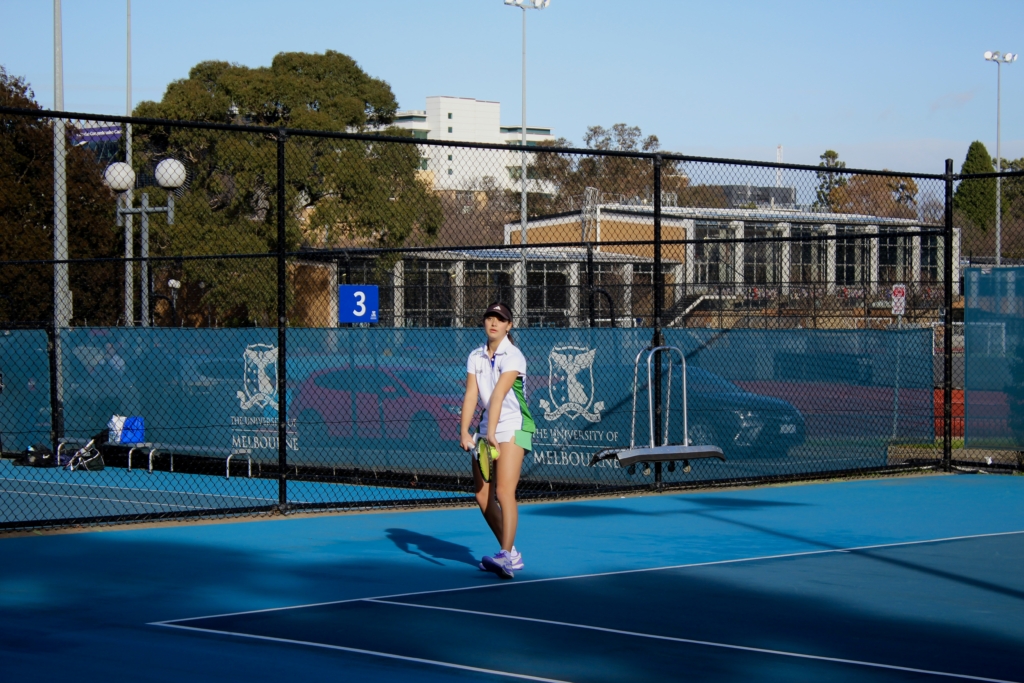 Female tennis player standing on a blue hard court near a chain-link fence, wearing a white and green outfit and cap, holding a racket.