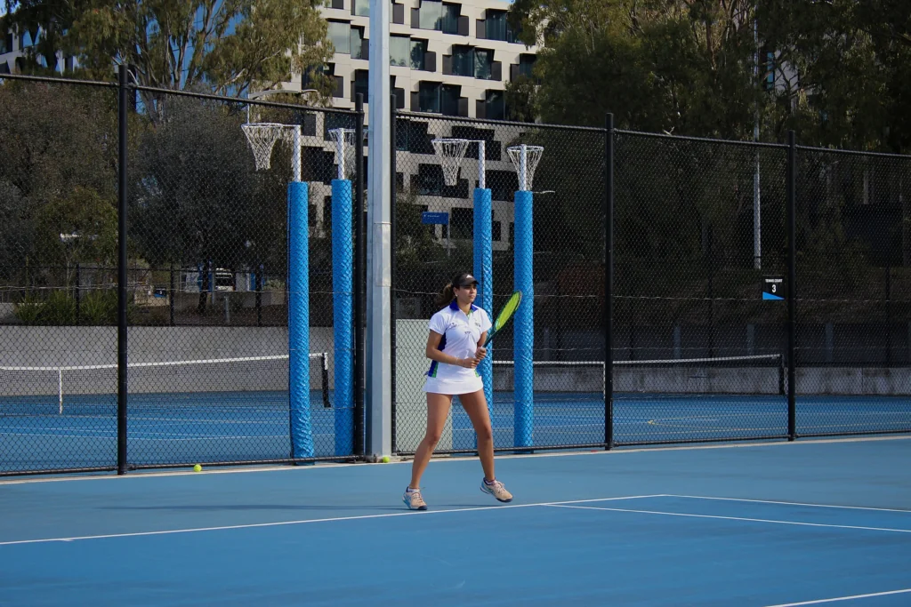 Female tennis player in white outfit standing on a blue outdoor court, behind a chain-link fence with blue padding on posts.