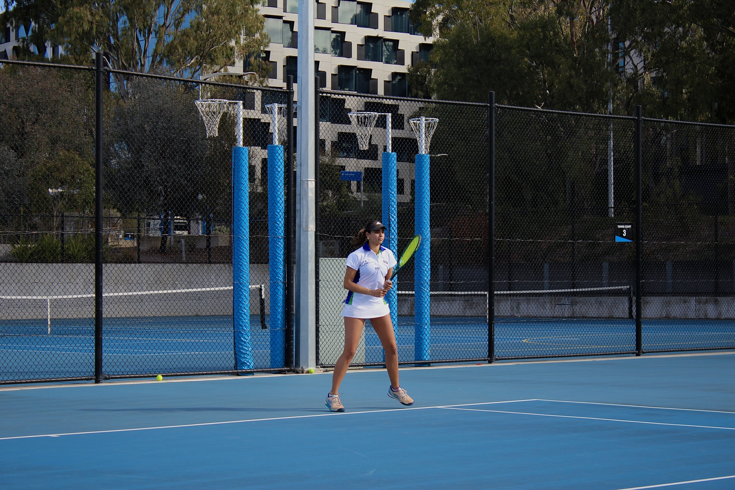 Female tennis player in white outfit standing on a blue outdoor court, behind a chain-link fence with blue padding on posts.