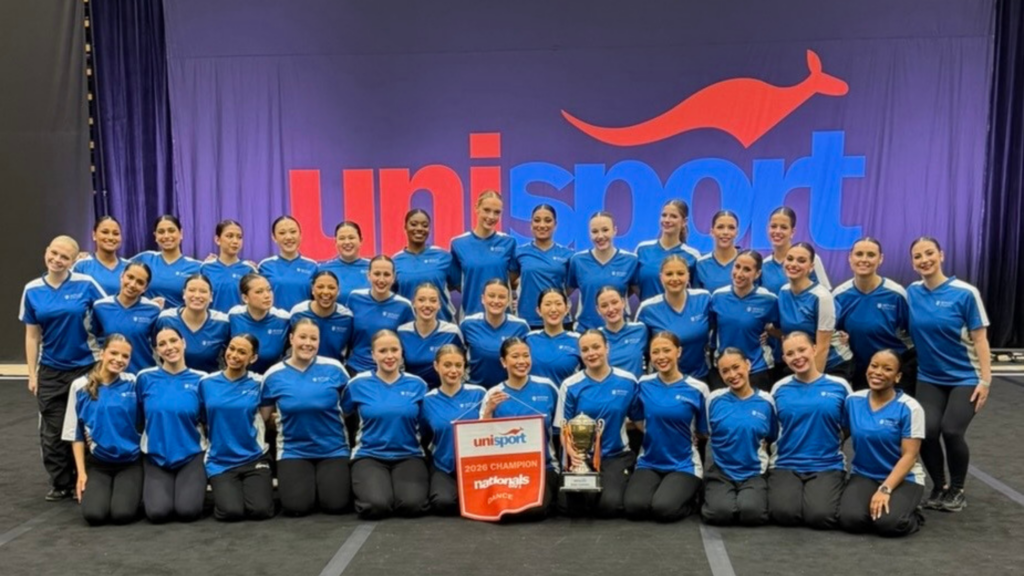 Team of athletes in matching blue shirts posing with trophy and banner in front of a red/blue UniSport backdrop.