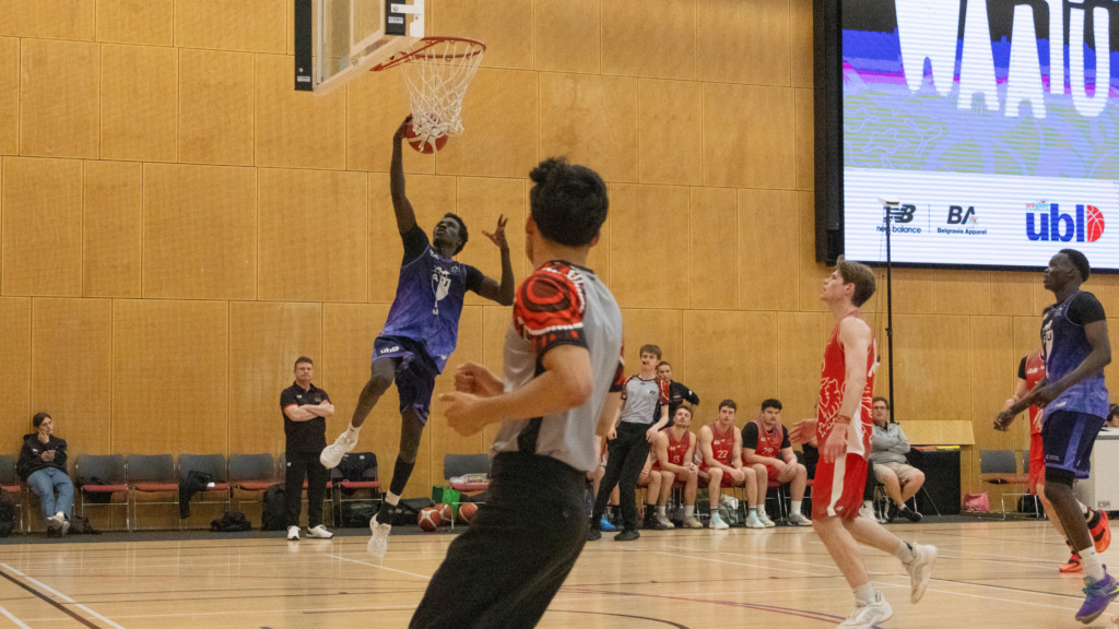 Player in blue jersey dunks toward the basket in a gym, with teammates and bench on the sideline watching.