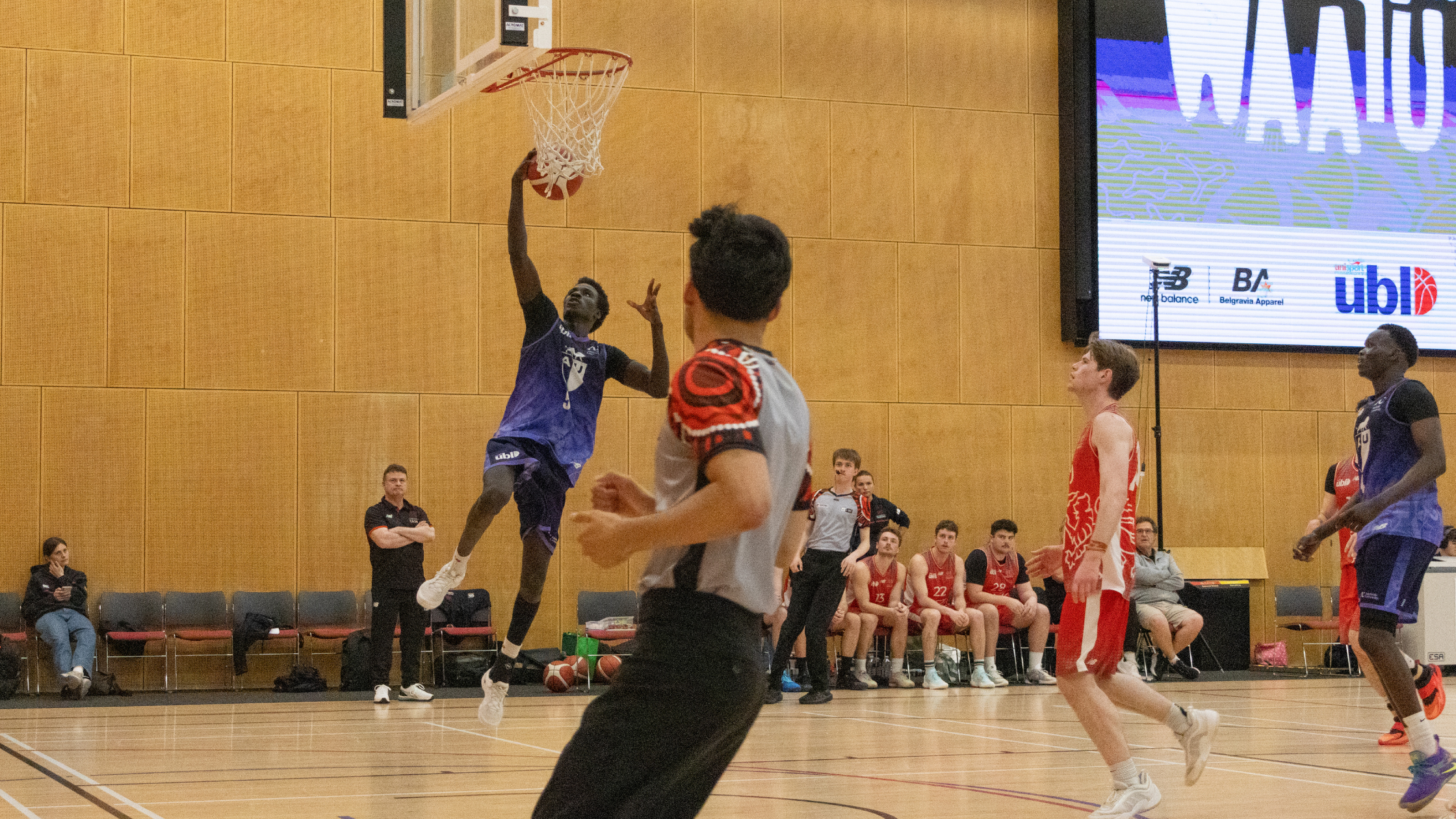 Player in blue jersey dunks toward the basket in a gym, with teammates and bench on the sideline watching.