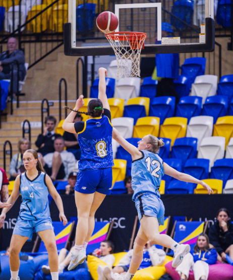 Two blue-uniform players jump near the basket as one goes for a layup while a defender reaches up to contest.