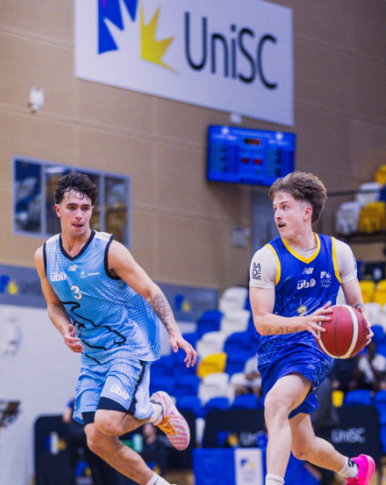 Two male basketball players sprint on an indoor court; player in blue dribbles the ball while a teammate or defender pursues, with a UniSC banner in the background.