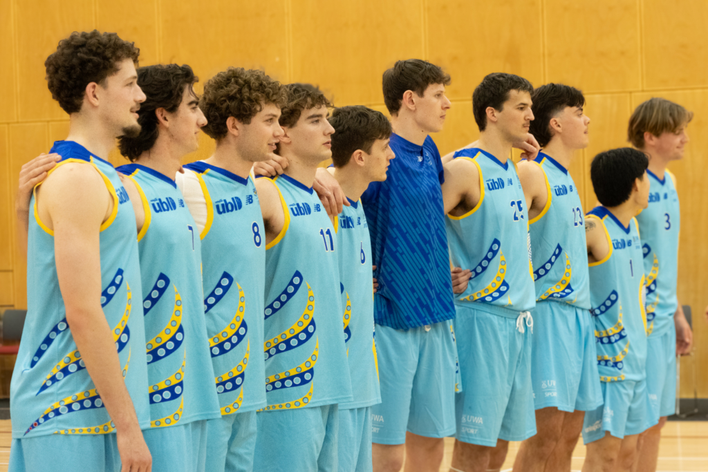 Line of male volleyball players in blue uniforms with yellow accents, standing shoulder to shoulder with arms around each other in a gym.