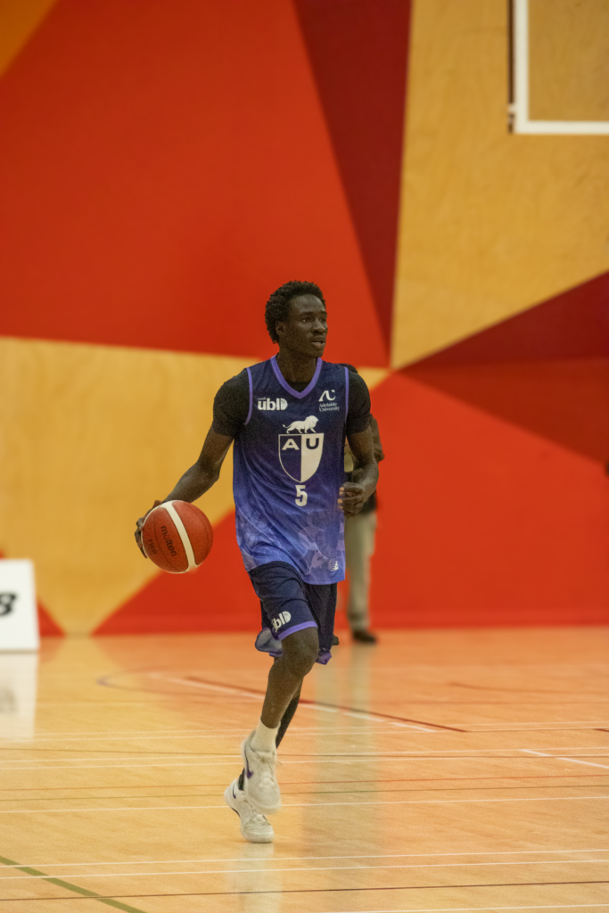 Basketball player in a blue uniform dribbles the ball on an indoor court with a bright orange and yellow geometric wall behind him.