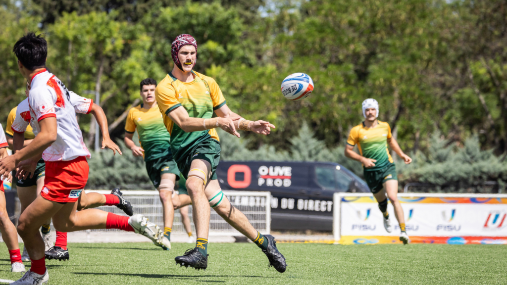 Rugby players chase a ball mid‑flight on a sunny field; foreground player in yellow‑green gears reaches for the ball while teammates and opponents rush nearby.