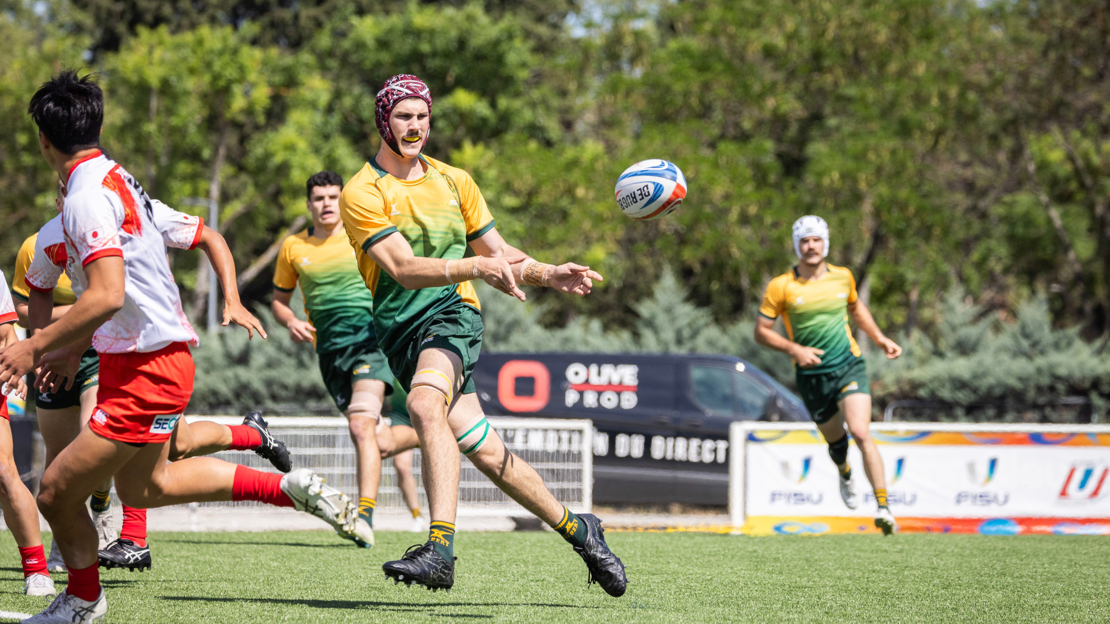 Rugby players chase a ball mid‑flight on a sunny field; foreground player in yellow‑green gears reaches for the ball while teammates and opponents rush nearby.