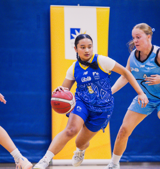 Female basketball player in a blue jersey (#5) dribbles the ball while being guarded by a defender in light blue on a gym court.